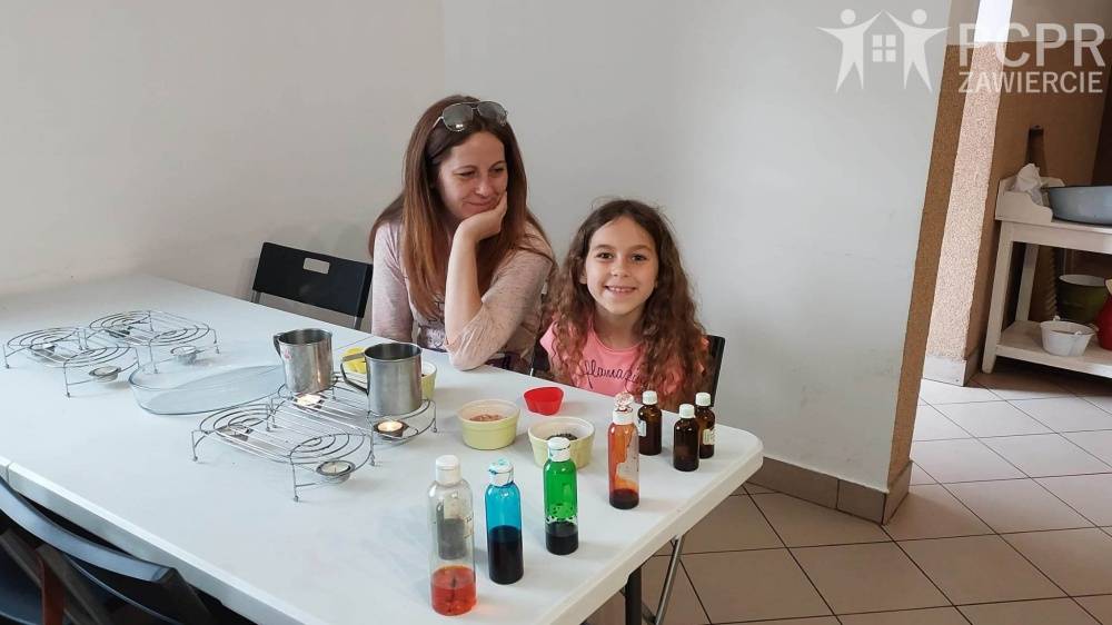 Zdjęcie: Uśmiechnięta kobieta i dziewczynka siedzą przy stole na warsztatach robienia świec. Przed nimi stoją garnuszki z woskiem i barwniki potrzebne do wykonania świec.